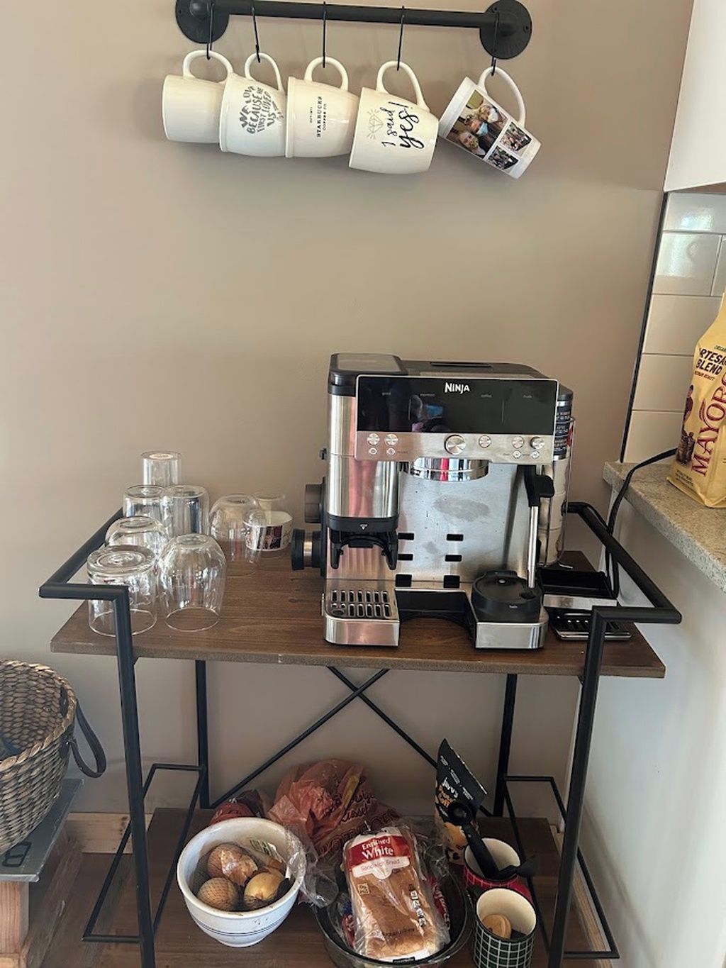 Una encimera de cocina con cafetera, tazas, vasos y alimentos.