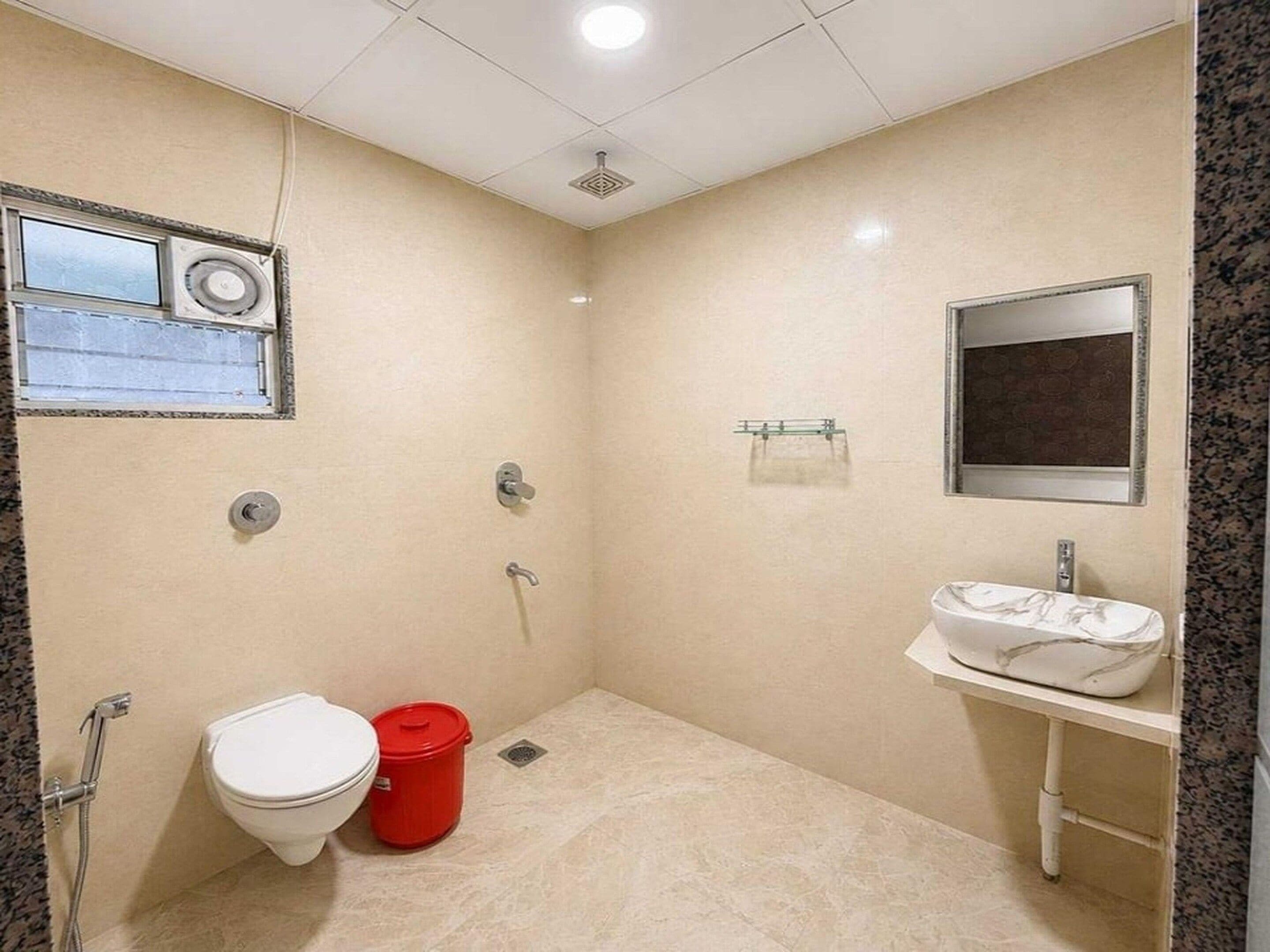 Un baño pequeño con inodoro, un cubo de basura rojo, lavamanos y espejo.