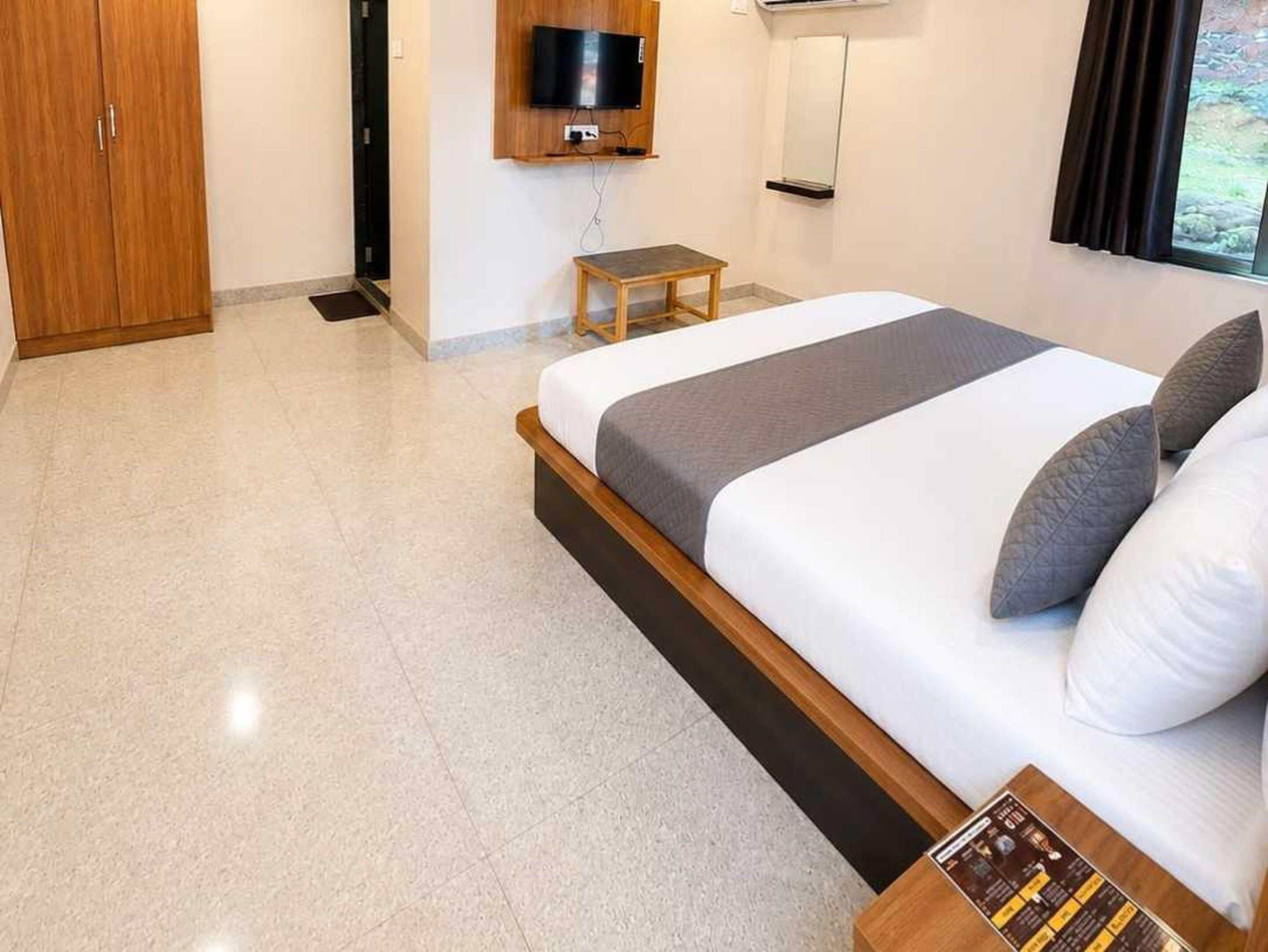 Ein Hotelzimmer mit einem Bett, einem an der Wand befestigten Fernseher, einem kleinen Tisch und einem Fenster mit Vorhängen.