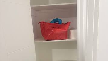 Ein schmaler Flur mit weißen Wänden und einer Tür rechts. Eine rote Tasche mit der Aufschrift 'FIRST AID' steht auf der zweiten Ablagefläche.