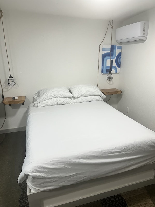 Un lit avec une literie blanche, un climatiseur fixé au mur et un luminaire suspendu.