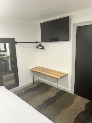 Een hotelkamer met een bed, een klein houten bankje, een kapstok, een televisie en een deur.