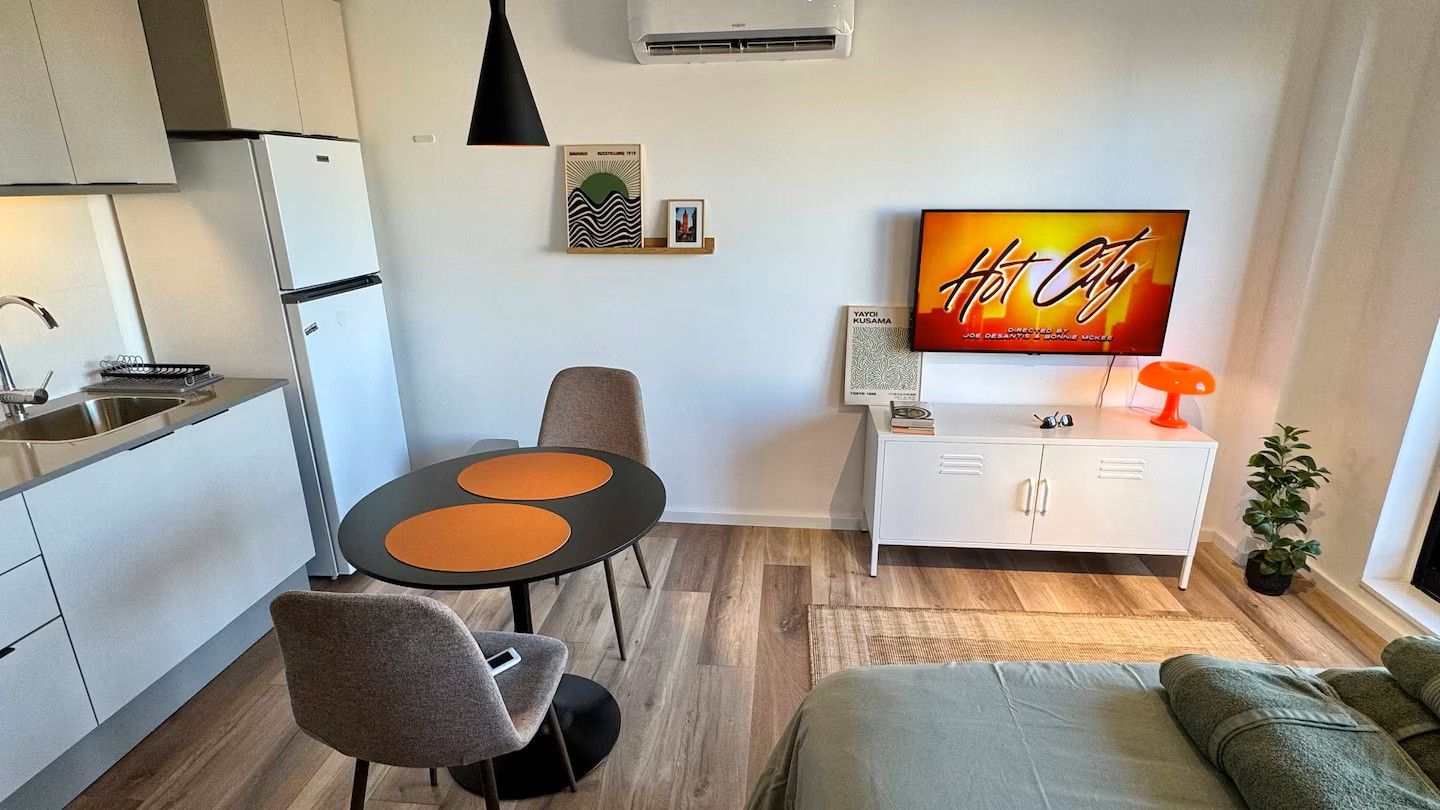 A compact living space with a kitchenette, a dining area with a table and chairs, a sofa, a TV mounted on the wall, and a potted plant.