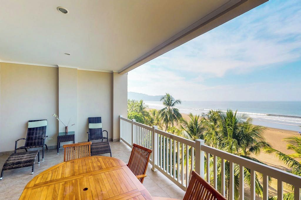 Un balcon avec une table et des chaises en bois, donnant sur une plage bordée de palmiers et sur l’océan.