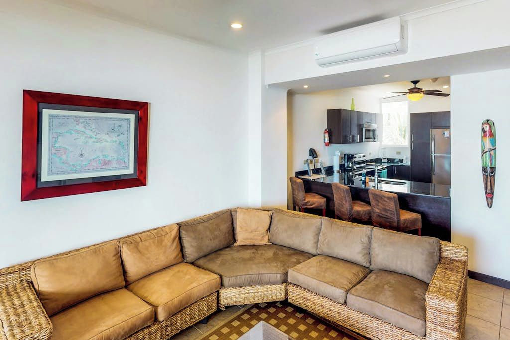 Uma sala de estar com um amplo sofá modular, uma área de jantar com cadeiras e uma cozinha visível por uma porta aberta.