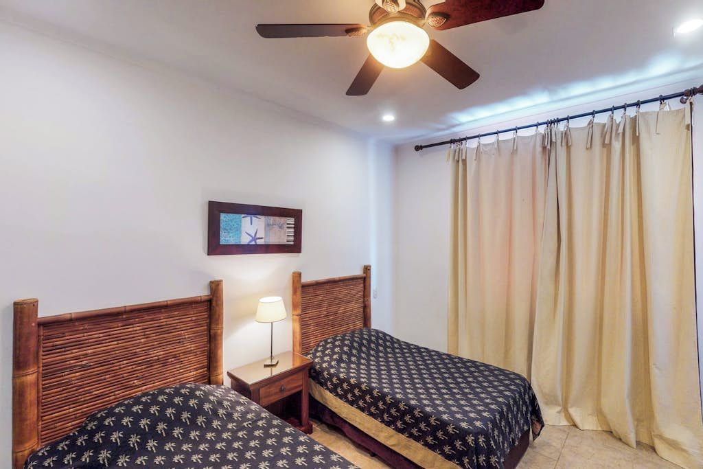 Una habitación con dos camas, un ventilador de techo, una mesita de noche con lámpara y una ventana con cortinas.