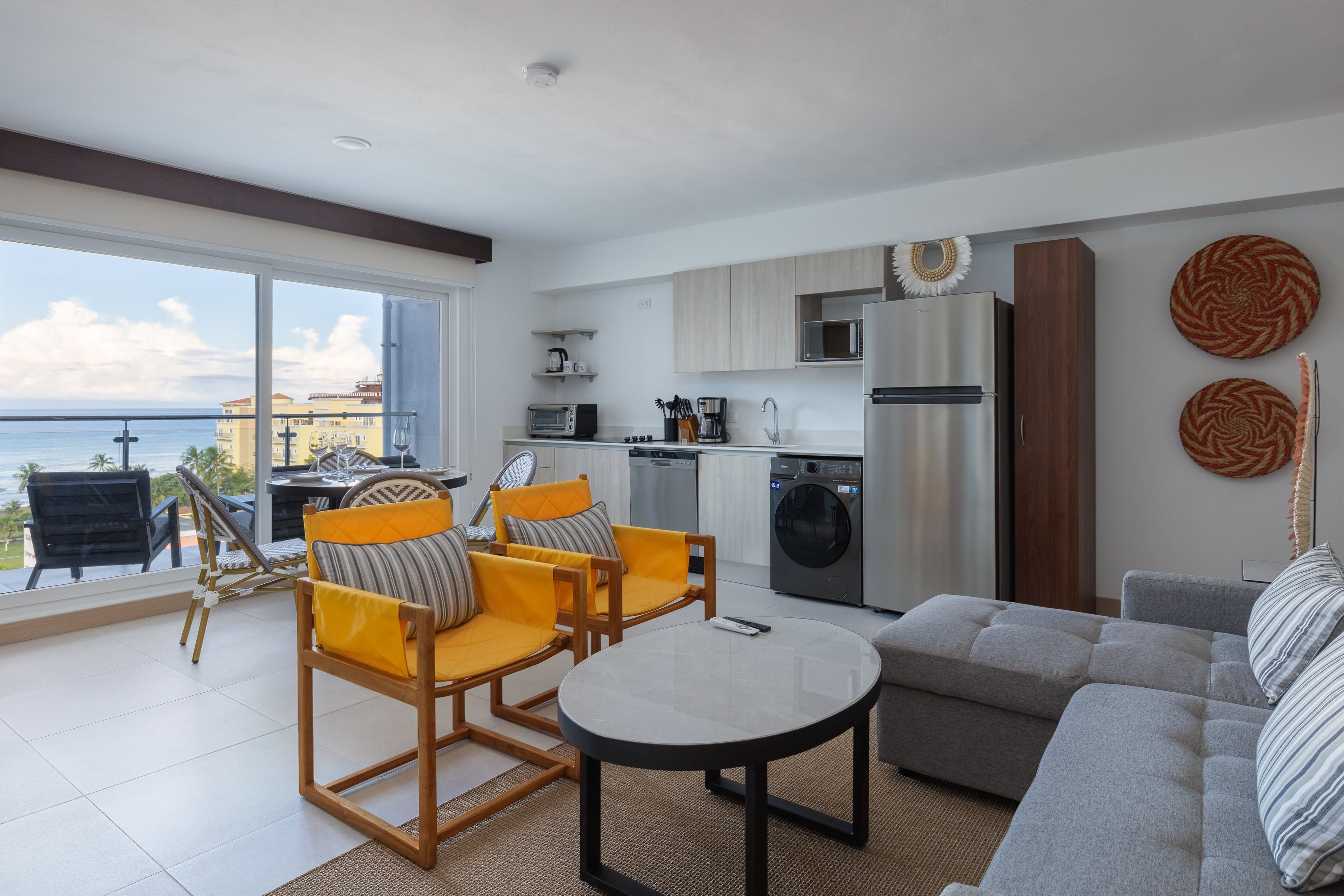 A modern living room with a sofa, chairs, and a coffee table. There is a kitchen area with appliances and a view of the sea.