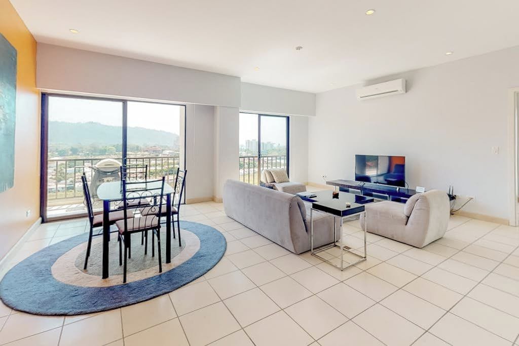 A modern living room with a dining area, a sofa set, a TV, and a balcony with a view.