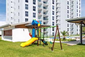 A playground with a slide and swings, a pool, and a building with balconies in the background. - Viva Jaco Large Capacity Condo (03-07C) (Jacó)