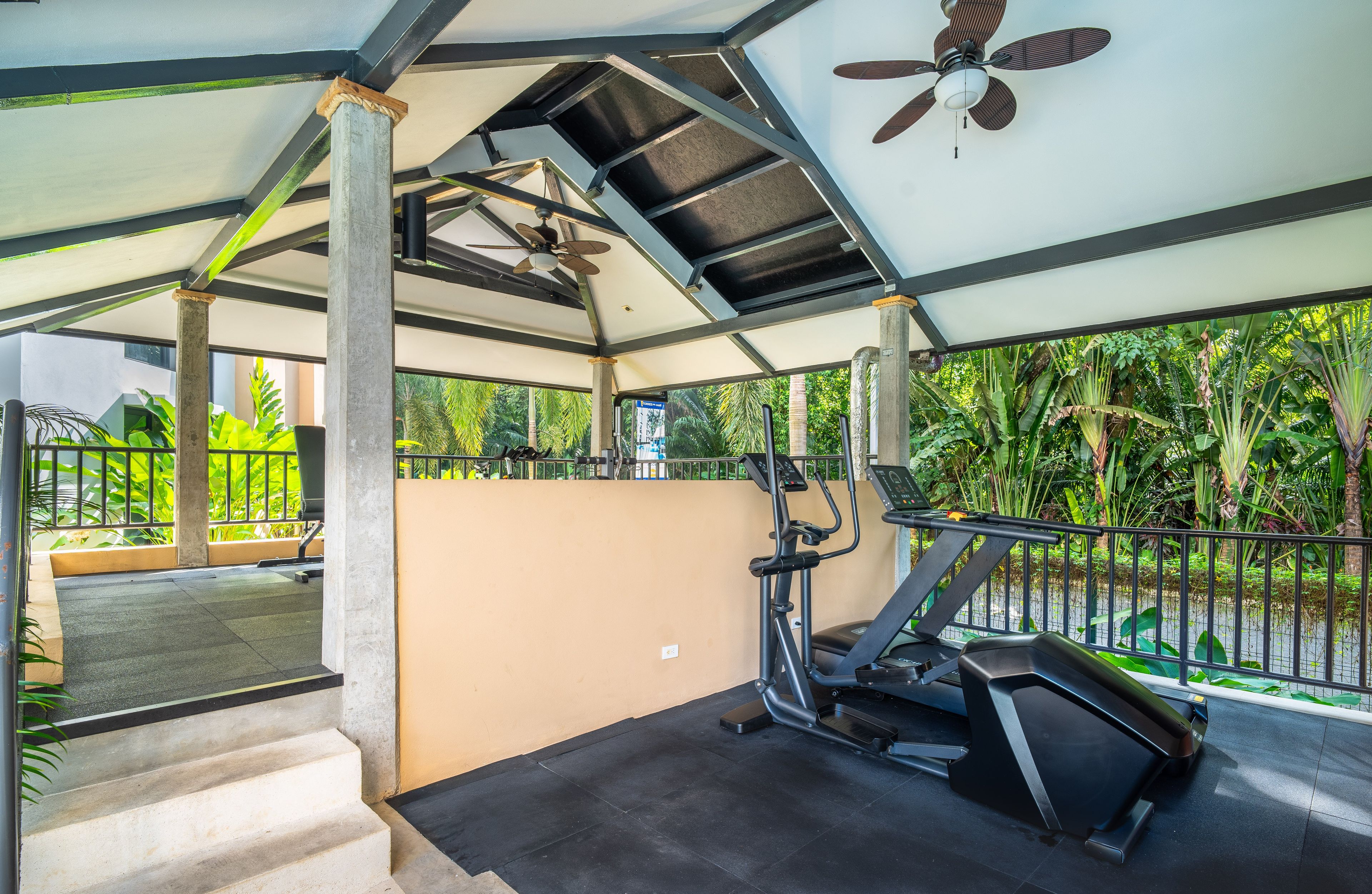 Een overdekte buitentrainingsruimte met fitnessapparatuur, een plafondventilator en een trap naar een balkon met uitzicht op weelderige begroeiing.