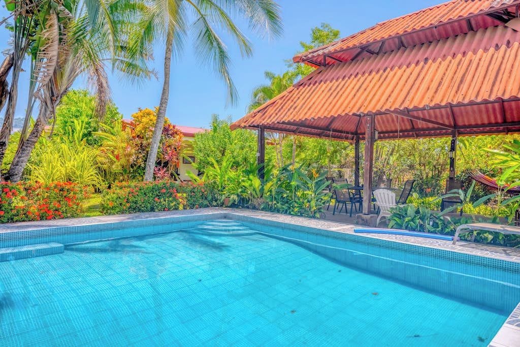 A swimming pool with a tiled edge, surrounded by lush greenery and a covered pavilion.