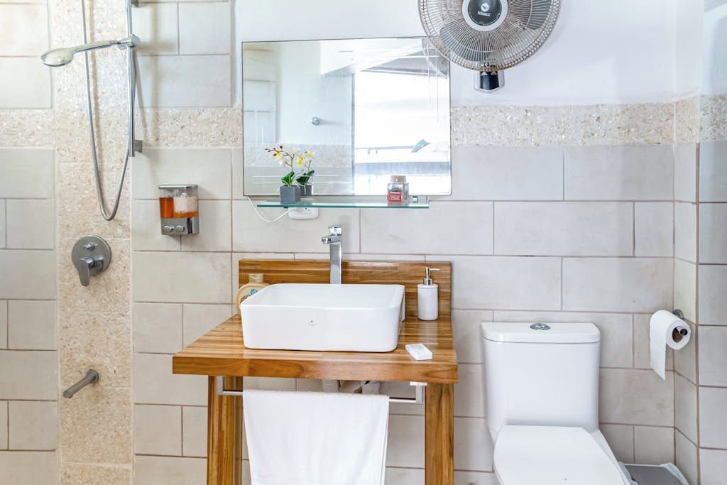 Une salle de bain avec un meuble-lavabo en bois, un lavabo blanc, des toilettes, une douchette à main, un ventilateur fixé au mur et un miroir.