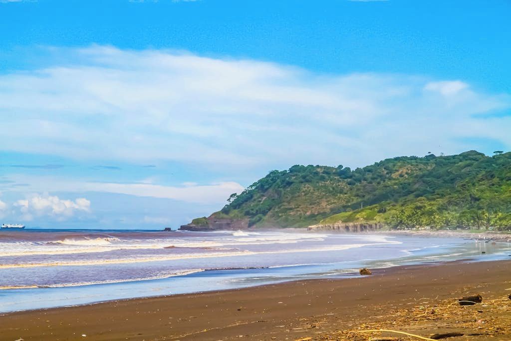 Une plage avec des vagues, un navire à l’horizon et une colline couverte de végétation.