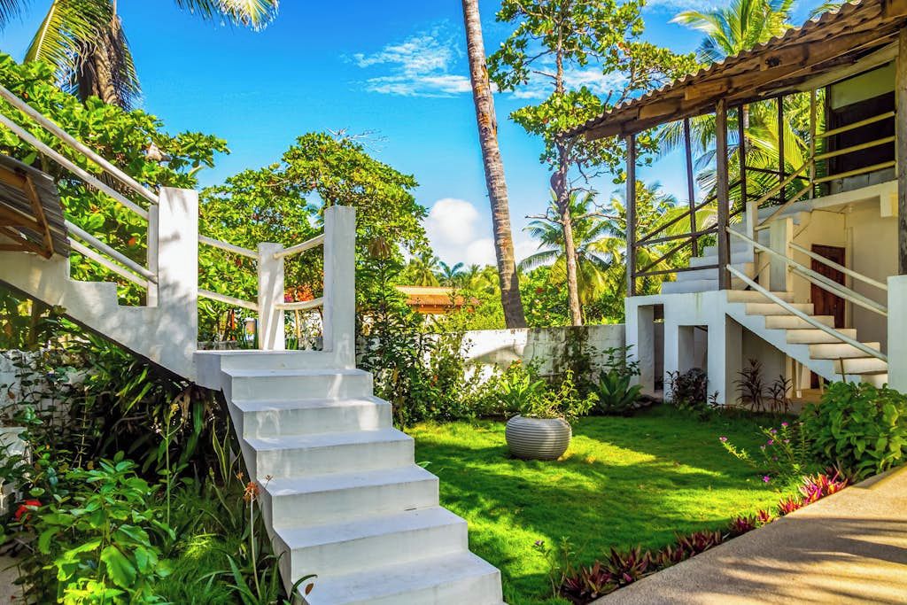 Un edificio moderno con una escalera que lleva a un área cubierta, rodeado de vegetación exuberante y palmeras.