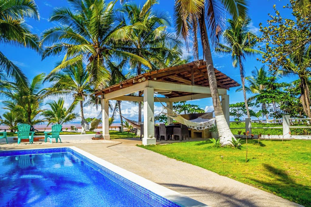 A pool area with a covered seating area, palm trees, and a clear sky.