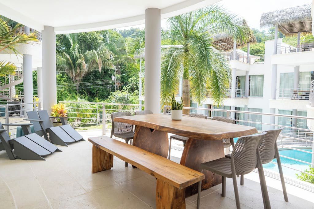 Un espace repas extérieur moderne, avec une table et des bancs en bois, entouré de chaises, et offrant une vue sur une piscine et une végétation luxuriante.