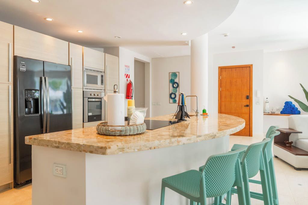 A modern kitchen with a central island, stainless steel appliances, and a seating area with green chairs.
