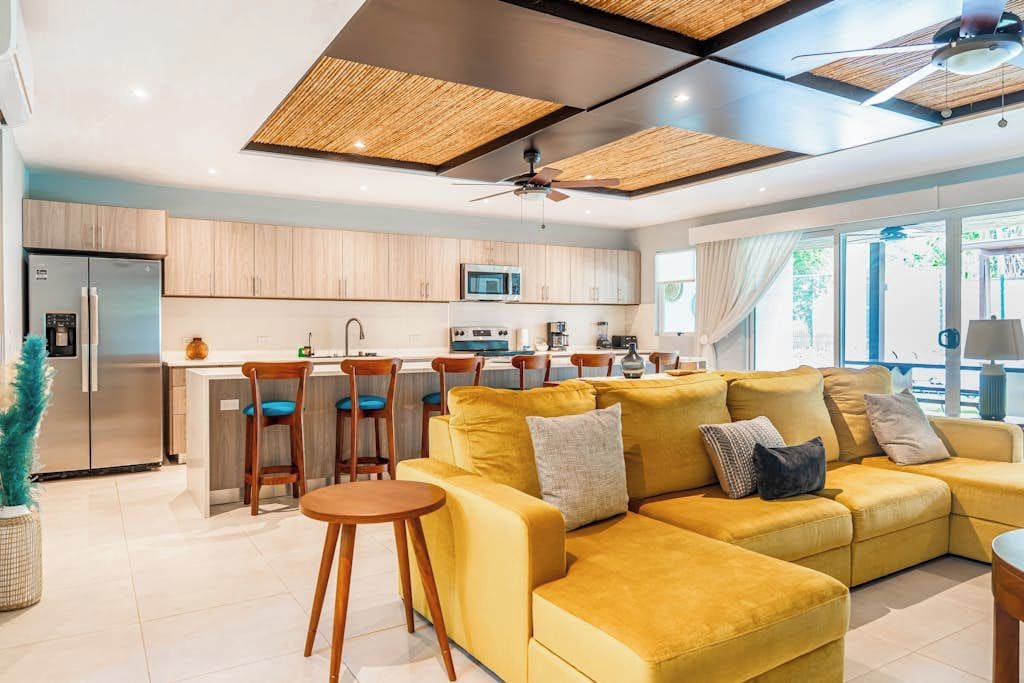 A modern kitchen with a central island, wooden cabinets, and a dining area with yellow sofas and chairs.
