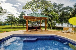 Una piscina con borde de azulejos, un pabellón de madera y sillas de descanso.