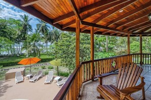 Un área de estar al aire libre con techo, mobiliario de madera y vistas a un jardín exuberante, con barandilla de madera.