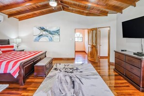 Un dormitorio con techo de madera, una cama con colcha a rayas rojas y blancas, un cuadro abstracto grande, un televisor de pantalla plana y suelo de madera.