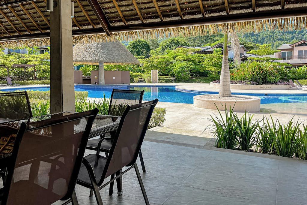 Área externa com assentos, vista para piscina, telhado de palha e paisagismo tropical.