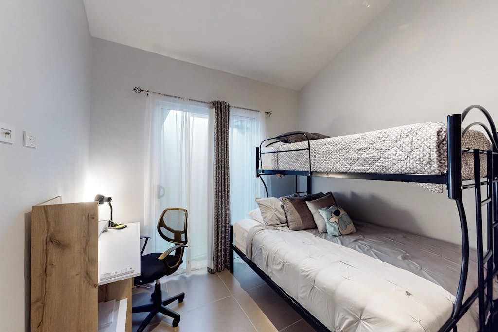 Een stapelbed met bureau en stoel, een raam met gordijnen en een houten bureau met lamp.