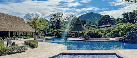 Ein Swimmingpool mit Wasserspiel, umgeben von Liegestühlen und einem Gebäude mit Reetdach.