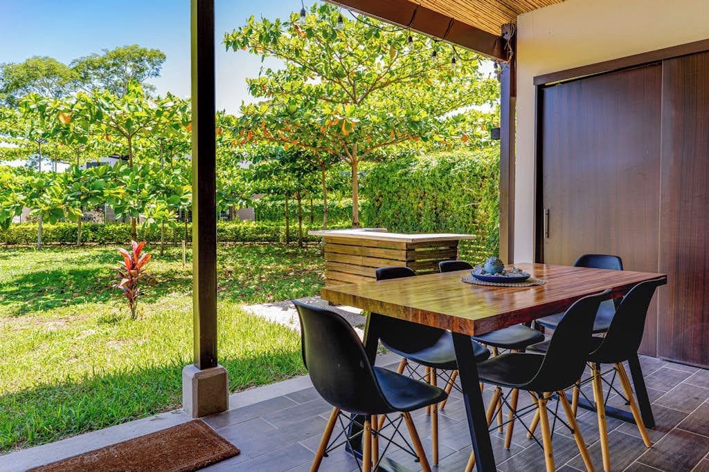 An outdoor dining area with a wooden table and chairs, surrounded by a garden with trees and grass.