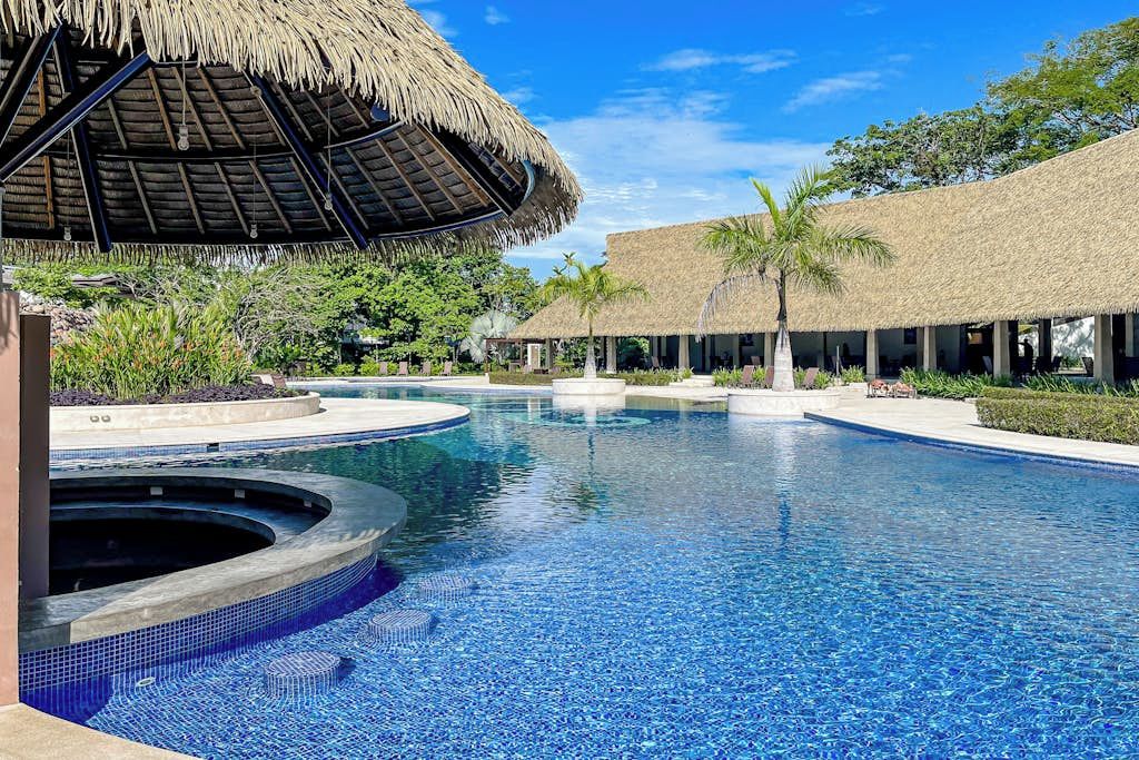 Uma piscina com área circular deofurôs, cercada por construções com telhado de palha e palmeiras.