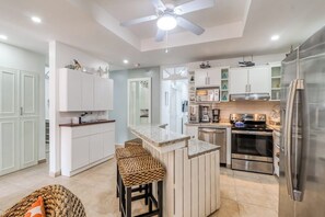A modern kitchen with white cabinetry, a central island with stools, stainless steel appliances, and a ceiling fan. - Bahia Encantada C2 Beachfront 3BR Ocean View Condo (Jaco)