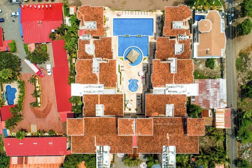 An aerial view of a hotel with multiple buildings, a central swimming pool, and surrounding greenery.