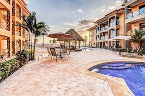 A hotel pool area with hexagonal tiles, lounge chairs, umbrellas, and a thatched roof structure. - Bahia Encantada C2 Beachfront 3BR Ocean View Condo (Jaco)