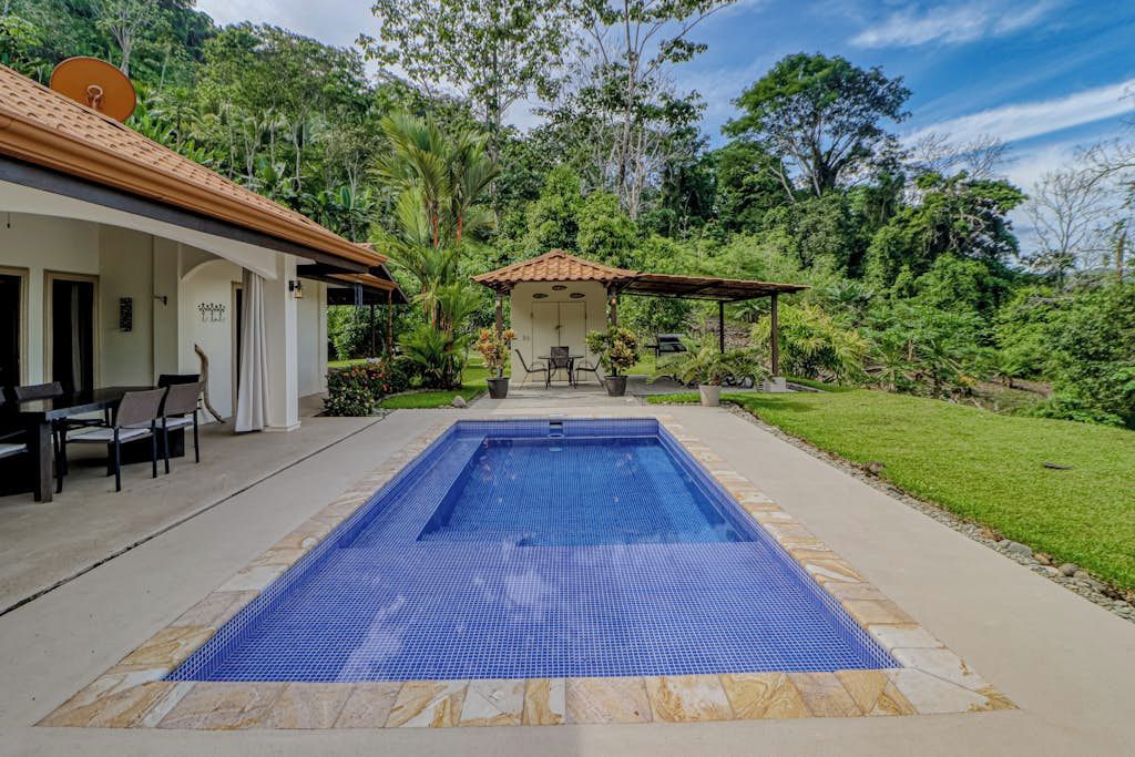 Una piscina con solárium revestido, un área de estar techada y un edificio con techo de tejas.