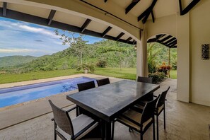 An outdoor patio with a pool, a table, and chairs, overlooking a lush landscape.