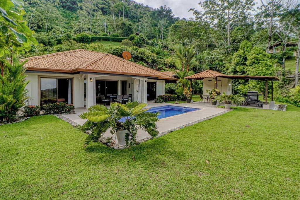 Una casa de dos pisos con piscina, rodeada de exuberante vegetación y colinas.