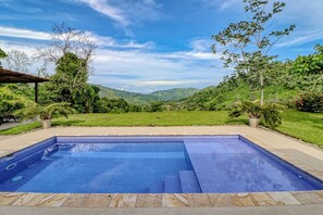 A rectangular swimming pool with a tiled edge, surrounded by a stone deck, set against a backdrop of lush greenery and distant hills.