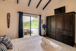 A bedroom with a bed, a dark wooden wardrobe, a television on the wall, and a view of a swimming pool outside.
