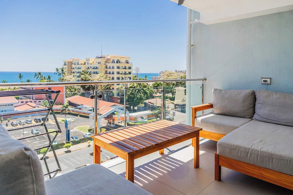 A balcony with a wooden table and a grey sofa, overlooking a cityscape and the sea.