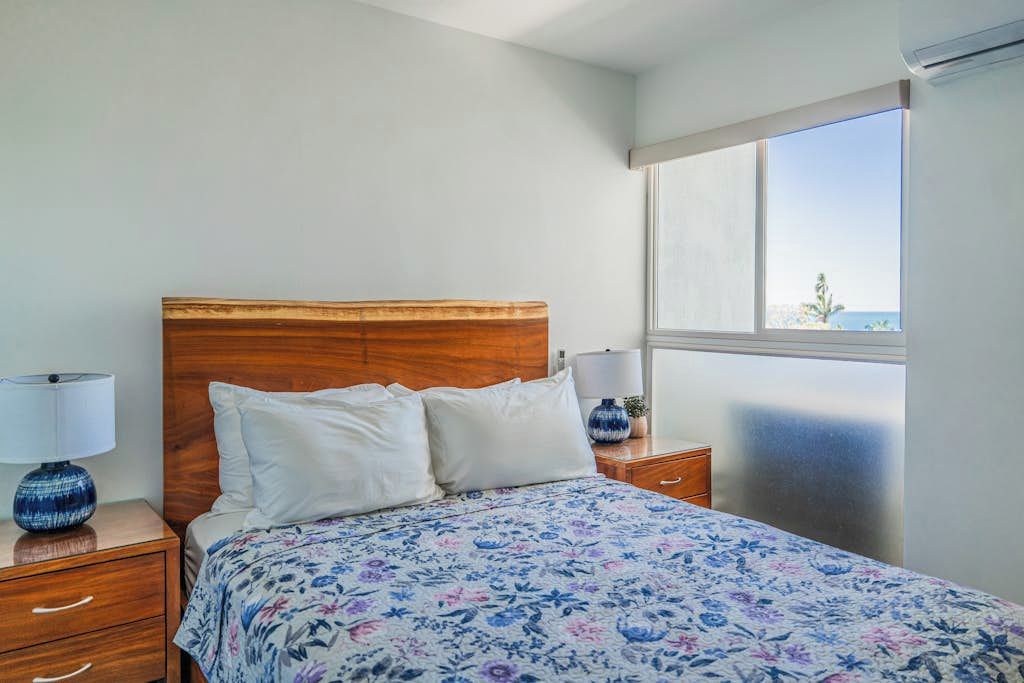 Una camera da letto con testiera in legno, copripiumino floreale, lampada da comodino e vista sul mare attraverso un'ampia finestra.