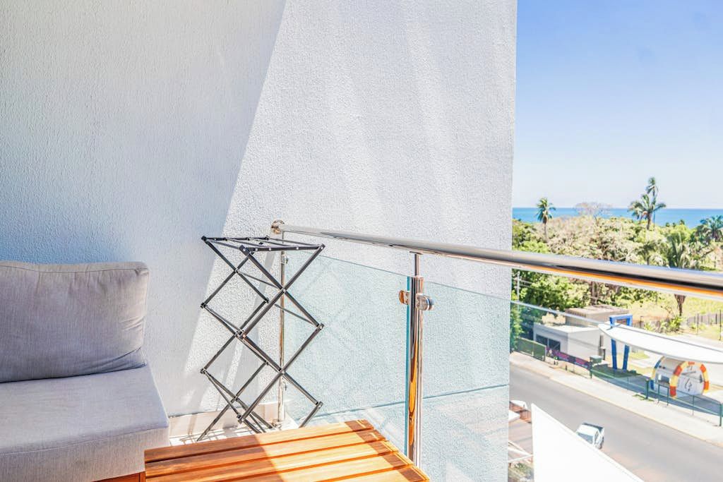 A balcony with a cushioned seating area, a metal drying rack, and a view of the ocean and a road with buildings.