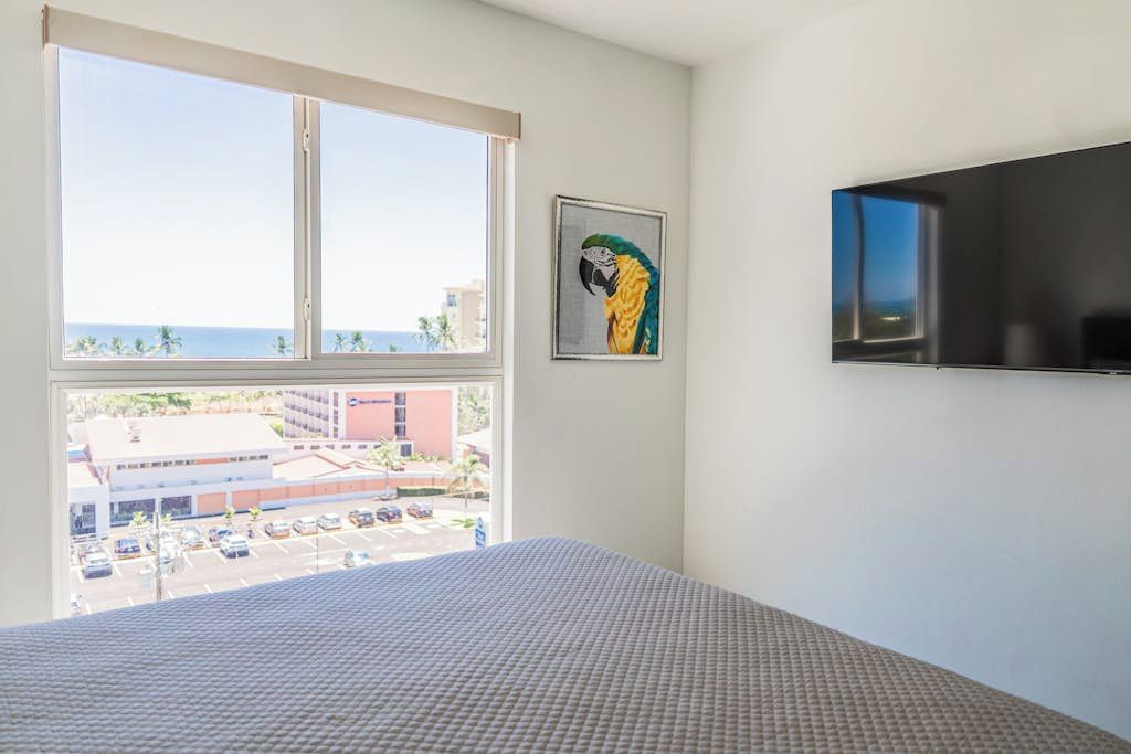 Ein Hotelzimmer mit großem Fenster, einem Bett, einem Flachbildfernseher und einem gerahmten Kunstwerk einer Papageien-Darstellung.