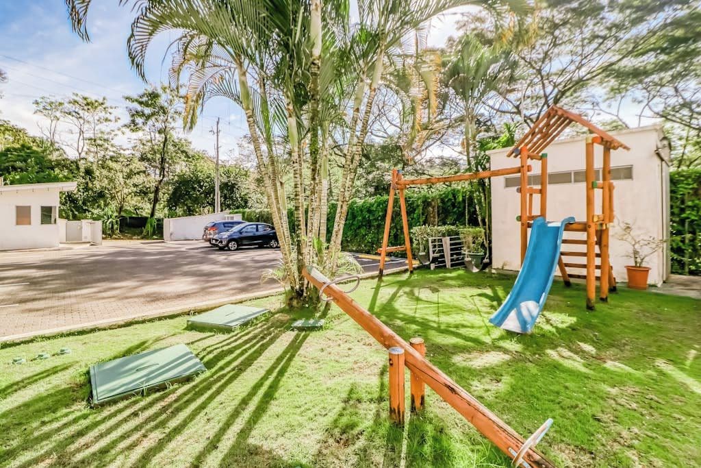 A residential area with a playground featuring a slide and swings, surrounded by greenery and a road.
