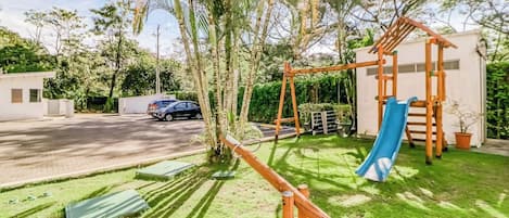 A residential area with a playground featuring a slide and swings, surrounded by greenery and a road.
