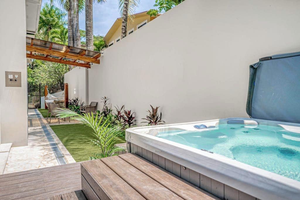 A hot tub in a backyard with a wooden deck, a pergola, and a small lawn area.