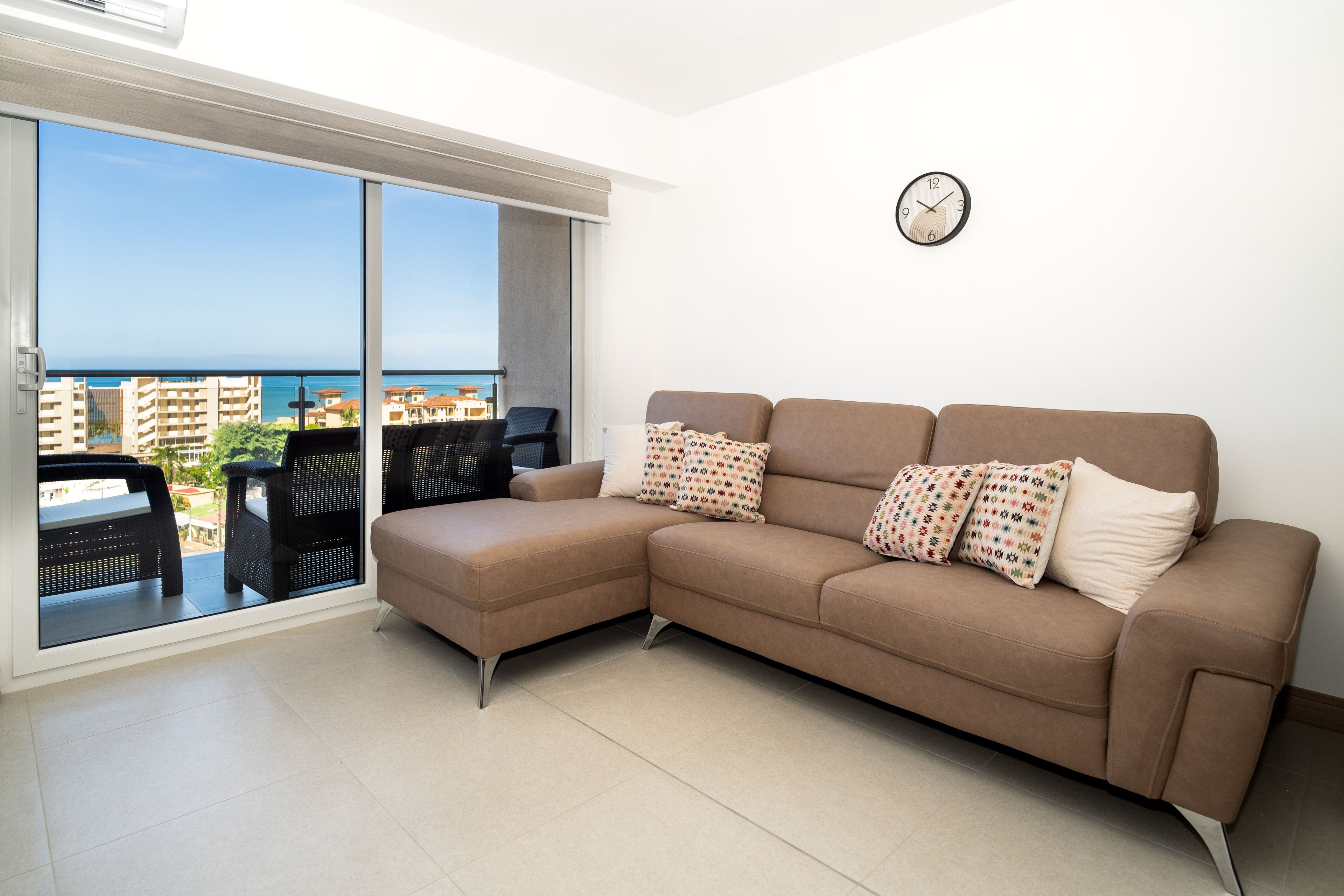 A modern living room with a brown sectional sofa, two patterned pillows, a wall clock, and a large sliding glass door leading to an outdoor balcony with a view of the sea and buildings.