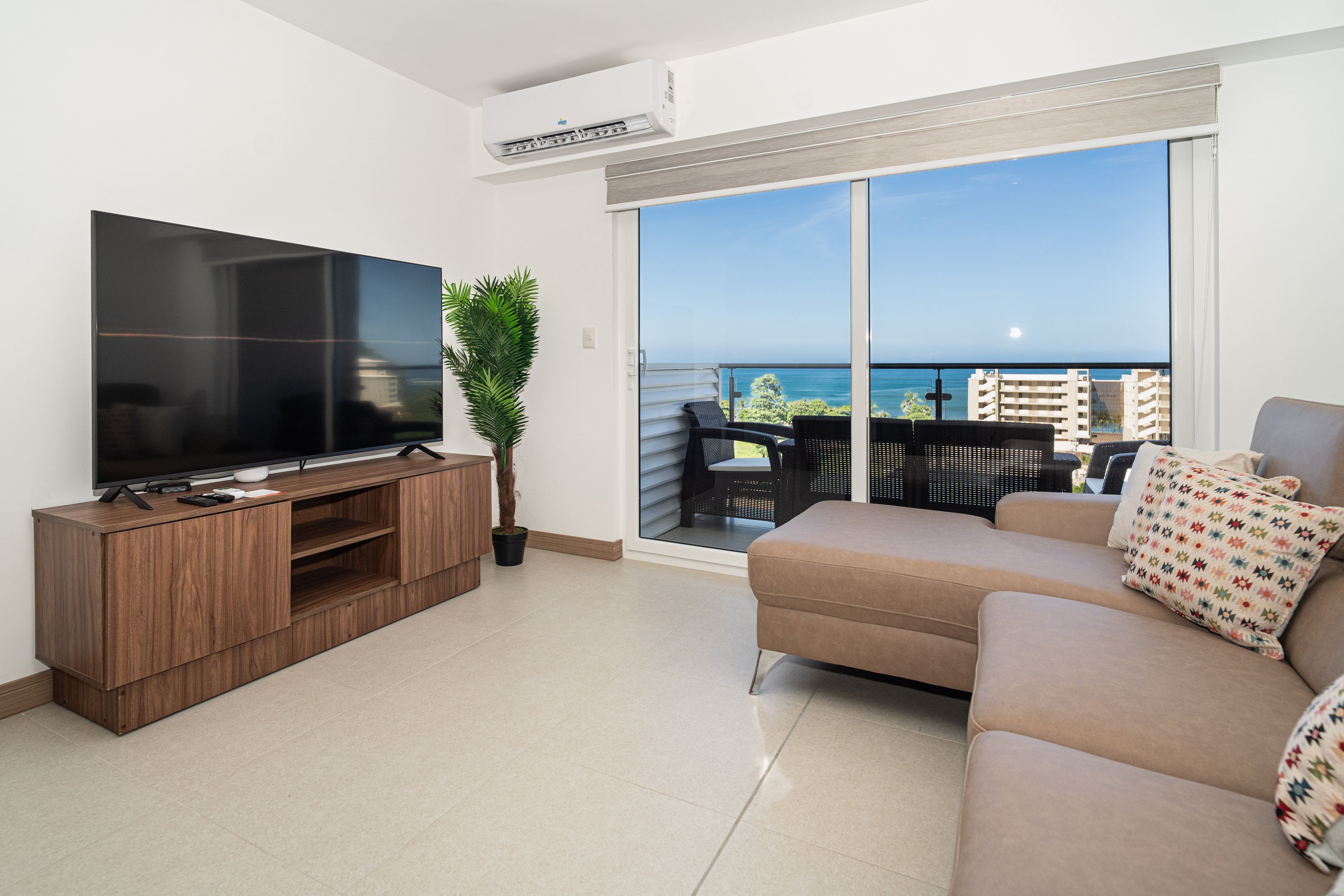 A modern living room with a flat-screen TV, a wooden entertainment unit, a potted plant, a beige sofa with patterned cushions, and a balcony with a view of the sea and buildings.