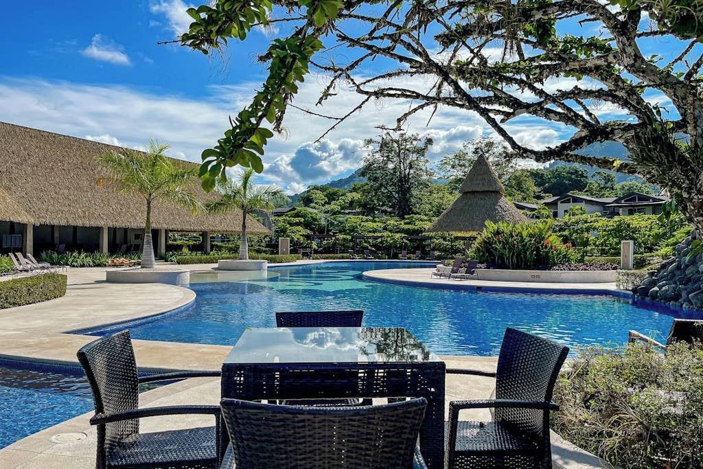 A swimming pool with lounge chairs and a table, surrounded by tropical vegetation and a mountain backdrop.