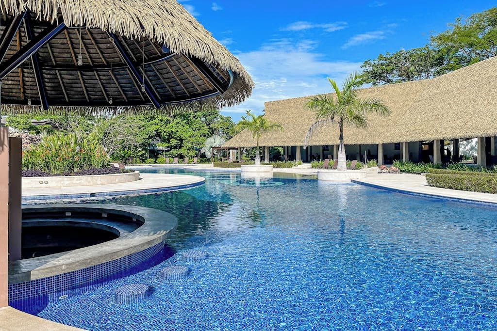 A swimming pool with a circular hot tub, surrounded by thatched-roof structures and palm trees.