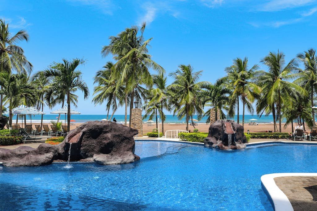 A pool with rock formations, palm trees, and a beach view.